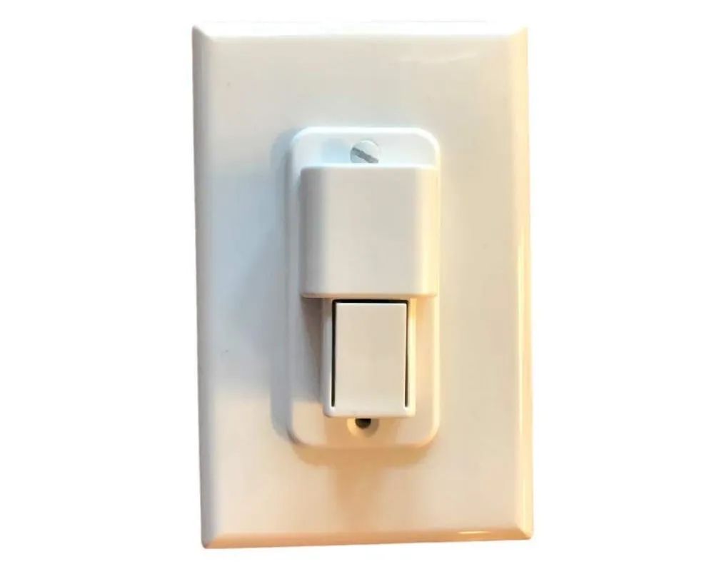 The white child-safety cover installed over a toggle switch on a standard wall plate, its button at the top and sliding carriage below.