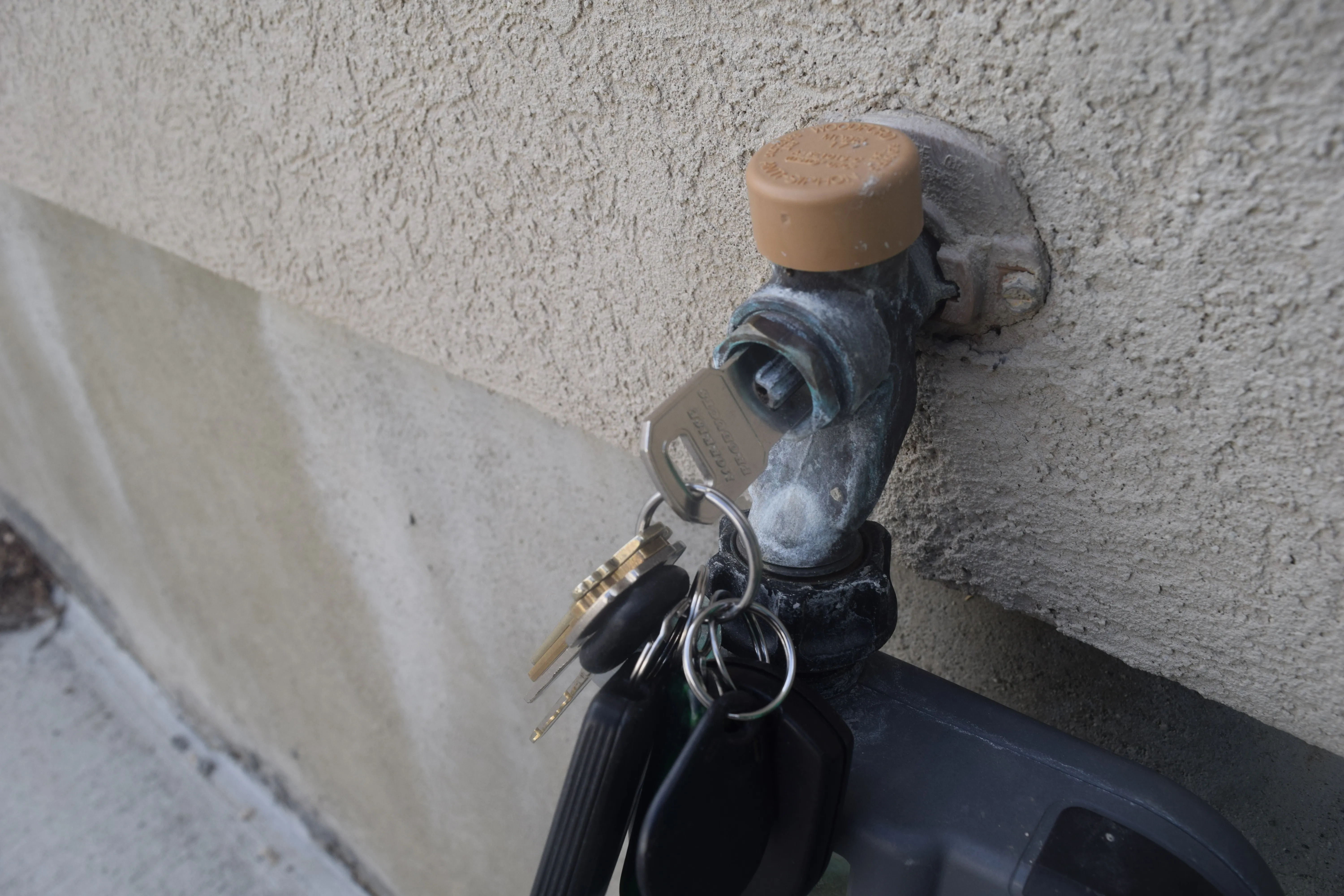 The tan replacement cap installed on an outdoor hose spigot against a stucco wall, with the steel key inserted into the stem and a loaded keyring hanging from it.